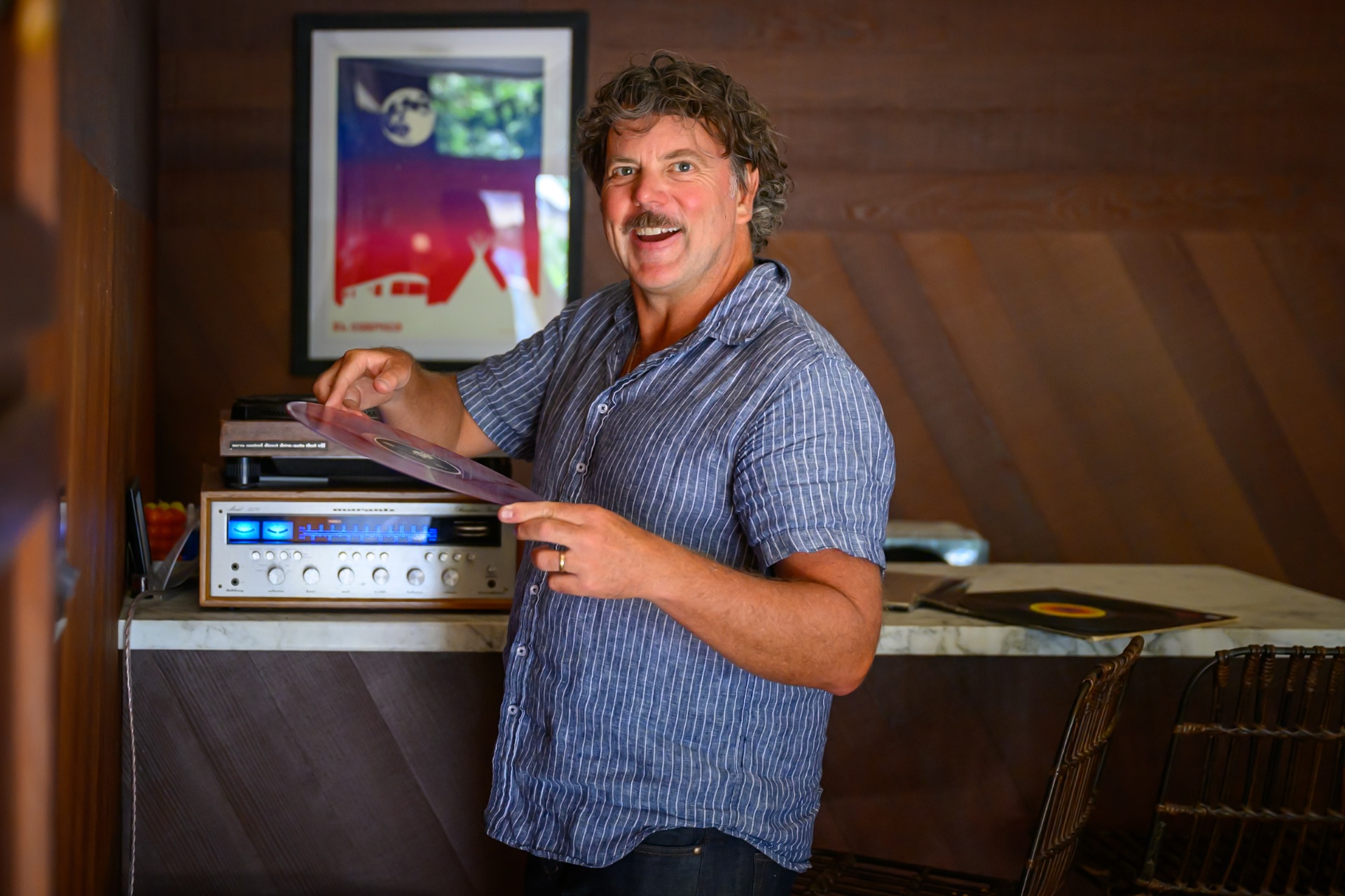 Chris holding a record beside a silver stereo in a wood-paneled room